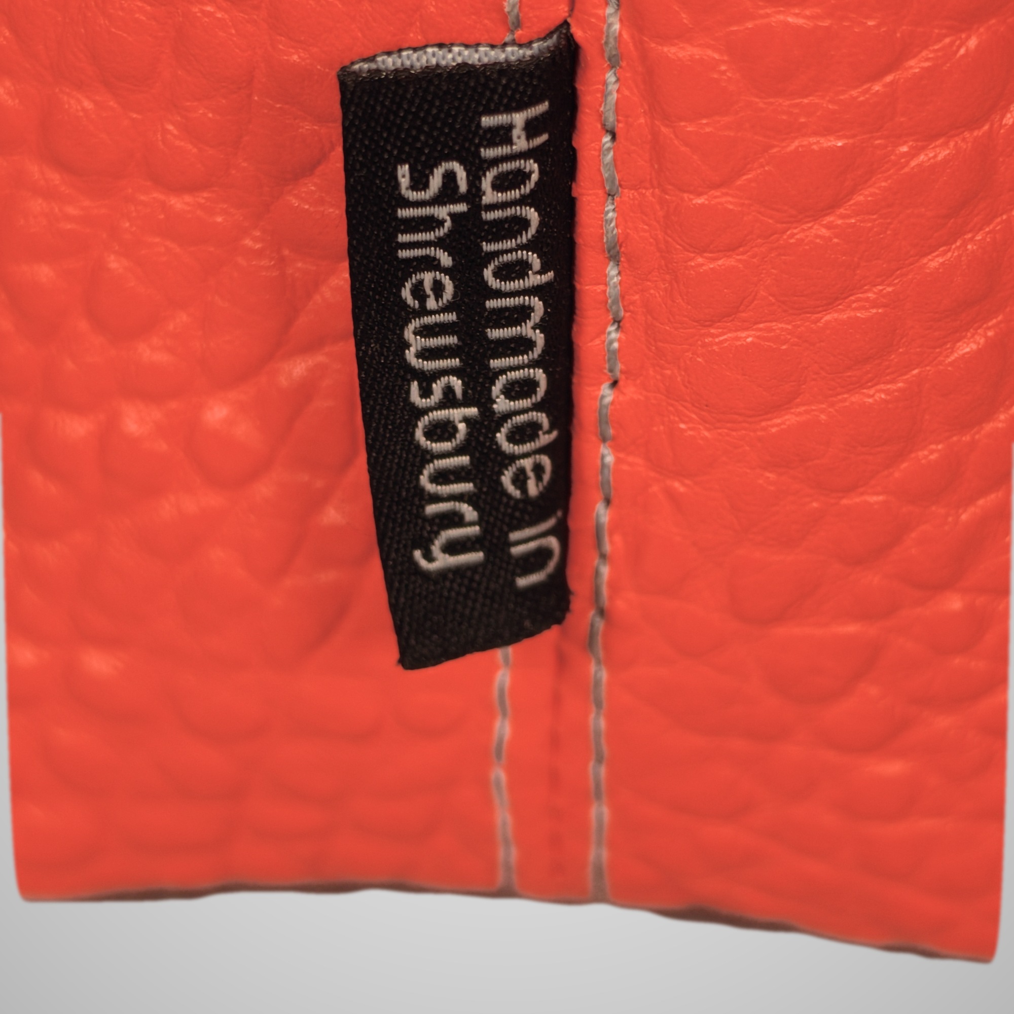Close-up of the “Handmade in Shrewsbury” label on the bright orange pebbled leather side seam